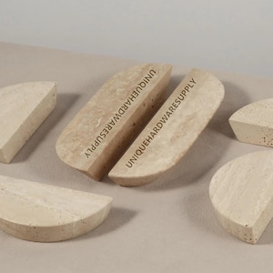May include: A selection of beige, semi-circular stone objects. Two larger pieces feature the text "UNIQUEHARDWARESUPPLY". The other pieces are smaller and of a similar shape. The objects are arranged on a neutral-coloured surface.
