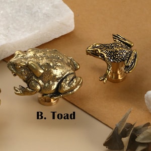 May include: Three brass frog and toad cabinet knobs. The knobs are detailed with textured surfaces and are mounted on a round base. The image includes the text "A. Frog" and "B. Toad".