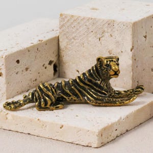 May include: A detailed, bronze-colored tiger figurine in a resting pose. The tiger is placed on a small, square, off-white stone. The background features two larger, off-white stone blocks and a burgundy flower.