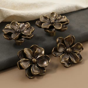 May include: Four bronze-colored flower-shaped cabinet knobs. The knobs are arranged on a dark gray surface, with a beige cloth in the background. The knobs have a detailed floral design, suitable for furniture hardware.