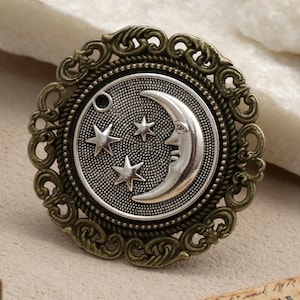 May include: A celestial-themed brooch with a silver crescent moon and three stars on a textured background. The brooch has an ornate, bronze-colored frame with a circular design. The brooch is a decorative accessory.