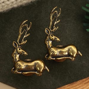 以下が含まれることがあります： 詳細な角と座ったポーズが特徴の、2つのアンティーク真鍮製の鹿の置物。 鹿は濃い緑色の表面にあり、金色のノブと緑色の小枝が背景にあります。 これらは装飾品である可能性が高いです。
