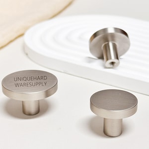 May include: Three brushed nickel cabinet knobs. One knob has the text "UNIQUEHARD WARESUPPLY" engraved on the face. The knobs have a cylindrical base and a flat, circular top. The hardware is displayed on a white surface.