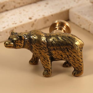 Puede incluir: Un tirador de armario con forma de oso de latón con un acabado texturizado. El oso está de pie sobre sus cuatro patas, con una representación detallada de su pelaje y sus rasgos. El tirador tiene una base cilíndrica para el montaje. El texto "UNIQUEHARD WARESUPPLY" es visible.