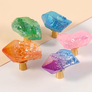 Peut inclure: Cinq boutons de meuble en cristal color&eacute; dans les tons de vert, orange, bleu, rose et violet. Chaque bouton a une base dor&eacute;e. Les cristaux ont un aspect rugueux et textur&eacute;.