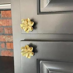 Op de afbeelding: Close-up van een deel van een donkergrijze kastdeur met twee goudkleurige bloemvormige kastknoppen. De onderste knop heeft de tekst "UNIQUE HARDWARE SUPPLY" erop. De kastdeur heeft een verzonken paneelontwerp.