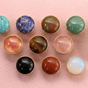 Può includere: Una collezione di pomelli decorativi per mobili con varie facce di pietre preziose, tra cui turchese, lapislazzuli e malachite. Ogni pomello ha un bordo in metallo dorato ed è posto su uno sfondo rosa.