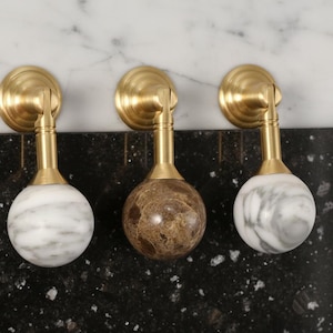May include: Three decorative cabinet knobs with brass hardware. Two knobs have white and gray marble spheres, and one has a brown and tan marble sphere. The knobs are mounted on a black surface.