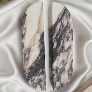 Op de afbeelding: Twee marmeren boekensteunen met een halfronde bovenkant en een rechthoekige basis. De boekensteunen zijn wit met donkerpaarse en zwarte adering. Ze staan tegen een witte satijnen achtergrond.