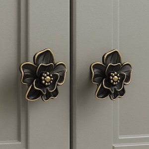 May include: Two black flower-shaped cabinet knobs with gold-colored edges and centers. The knobs are mounted on a light gray cabinet door. The floral design adds a decorative touch to furniture.