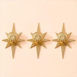 May include: Three gold-colored metal cabinet knobs, each featuring a central circular fluted knob mounted onto a flat, star-shaped backplate with eight pointed rays of varying lengths, creating a symmetrical and geometric hardware design.