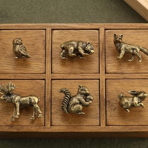 Puede incluir: Una caja de madera con seis pequeños cajones, cada uno adornado con una detallada figura de animal de latón. La colección incluye un búho, un oso, un zorro, un alce, una ardilla y un conejo, todos de estilo rústico.