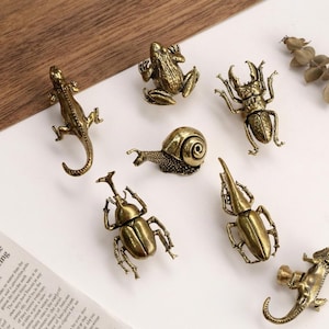 May include: A collection of small, antique brass animal figurines, including a lizard, frog, snail, and various beetles. The detailed sculptures are arranged on a white surface, with a newspaper clipping in the corner.