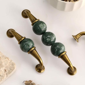 Op de afbeelding: Drie kastgrepen met antiek messingkleurig metalen beslag en ronde groene marmeren accenten. De grepen hebben gebogen metalen uiteinden en een centraal ontwerp met een of meer gepolijste groene stenen bollen met witte aders.