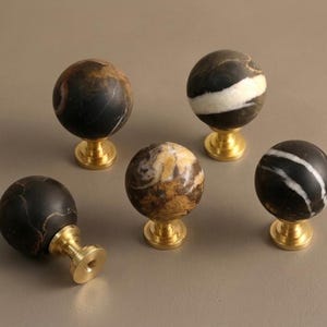 May include: Five spherical cabinet knobs featuring polished brass bases and natural stone tops. The stone spheres display varied patterns in black, white, and brown tones, with one knob lying on its side to reveal the circular brass mounting plate.