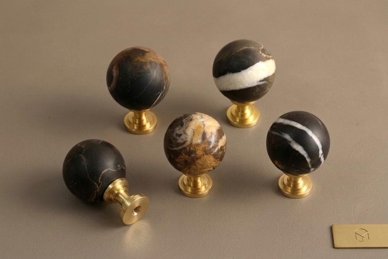 May include: Five spherical cabinet knobs featuring polished brass bases and natural stone tops. The stone spheres display varied patterns in black, white, and brown tones, with one knob lying on its side to reveal the circular brass mounting plate.