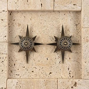 May include: Two identical star-shaped metal ornaments with a dark bronze finish. Each piece features a central circular medallion with a radiating petal pattern and a small raised dot in the middle, surrounded by eight sharp, pointed rays.