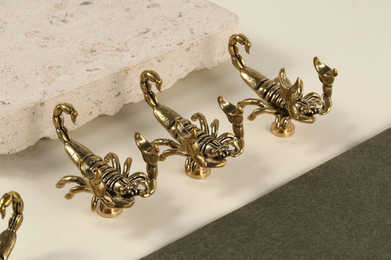 May include: Close-up of several brass scorpion-shaped cabinet knobs. The knobs are detailed with claws, segmented bodies, and curved tails. They are arranged on a white surface with a textured stone element.