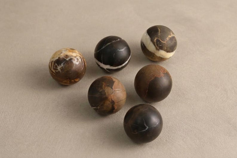 May include: A collection of six smooth, spherical stone objects in varying shades of dark brown, tan, and black. Each sphere features unique natural veining, swirls, and color patterns, with some displaying distinct white or cream-colored mineral streaks.