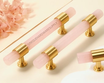 May include: Two long drawer pulls and one short drawer pull featuring cylindrical light pink translucent bars with gold-toned metal mounts at each end. The text UNIQUEHARDWARESUPPLY is printed in black on the surface of the longest drawer pull.