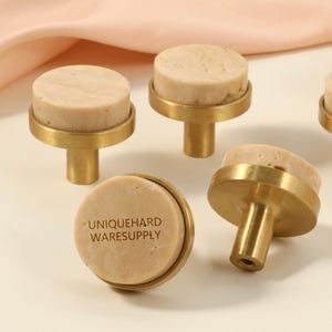 May include: Close-up of several cabinet knobs. Each knob features a light beige, circular stone top set on a brass base. The text "UNIQUEHARD WARESUPPLY" is printed on one knob. The knobs are designed for furniture.