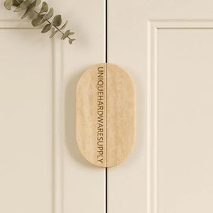 May include: A light beige, oval-shaped cabinet pull with the text "UNIQUEHARDWARESUPPLY" printed vertically. The pull is mounted on a white cabinet door. A sprig of greenery is visible in the upper left corner.