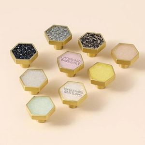 May include: A collection of decorative cabinet knobs with hexagonal faces in various colors, including black, white, pink, yellow, and green. Each knob has a gold-colored base and the text "UNIQUEHARD WARESUPPLY" printed on some of the knobs.