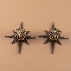 May include: Two antique brass cabinet knobs, each featuring a central fluted, circular knob mounted onto a flat, eight-pointed star-shaped backplate. The metal has a dark, brushed finish with visible highlights on the ridges of the fluted knobs.