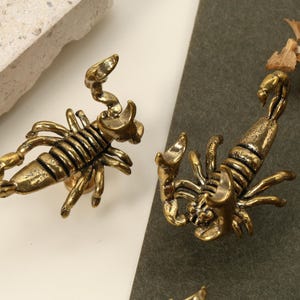 May include: Close-up of several antique gold-colored scorpion-shaped decorative objects. The scorpions are detailed with segmented bodies, claws, and curved tails. The objects are on a neutral background.