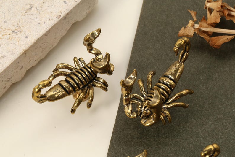 May include: Close-up of several antique gold-colored scorpion-shaped decorative objects. The scorpions are detailed with segmented bodies, claws, and curved tails. The objects are on a neutral background.