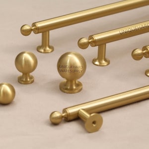 Może przedstawiać: Kolekcja złotych okuć do szafek, w tym gałki i uchwyty prętowe. Okucia mają gładkie, matowe wykończenie i prosty, nowoczesny design. Uchwyty prętowe mają różne długości. Tekst "UNIQUEHARDWARESUPPLY" jest wydrukowany.