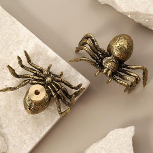 May include: Two antique brass spider figurines. The spiders have detailed legs and bodies, with a golden finish. One spider has a hole in its abdomen. The spiders are placed on a white marble surface.