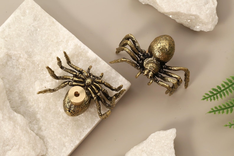 May include: Two antique brass spider figurines. The spiders have detailed legs and bodies, with a golden finish. One spider has a hole in its abdomen. The spiders are placed on a white marble surface.