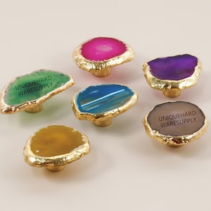 May include: Six decorative cabinet knobs with natural agate stone faces in shades of green, pink, purple, blue, brown, and yellow. Each knob has a gold-colored metal base and rim. The text "UNIQUEHARD WARESUPPLY" is printed on some of the knobs.