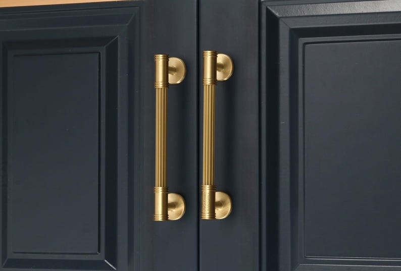 May include: Two vertical brass cabinet pulls with a ribbed, cylindrical design and circular mounting bases. The handles feature a brushed gold finish and are installed on dark navy blue cabinet doors with a recessed panel frame detail.