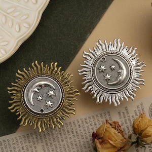 Puede incluir: Primer plano de tiradores decorativos para armarios con diseños celestiales. Los tiradores presentan un marco de rayos de sol alrededor de una luna creciente y estrellas. Un tirador es dorado, el otro plateado. Los tiradores están sobre un fondo neutro.