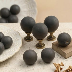 May include: A collection of spherical cabinet knobs featuring dark charcoal grey matte balls mounted on antique brass-toned metal bases. Several knobs are displayed upright on their bases, while others are shown as loose spheres.