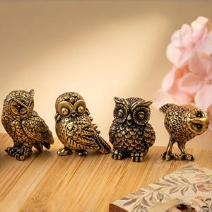 May include: Four detailed brass owl figurines in various poses are displayed on a wooden surface. The owls have intricate feather detailing and a metallic finish. A pink floral arrangement is visible in the background.