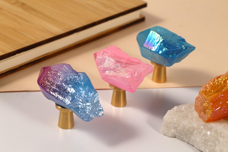 Peut inclure: Gros plan sur des boutons de placard en cristal color&eacute;. Les boutons sont dans des tons de bleu, de rose et d'orange, avec un aspect cristallin textur&eacute;. Chaque bouton est mont&eacute; sur une petite base dor&eacute;e. Le texte "UNIQUEHARD WARESUPPLY" est visible sur l'un des boutons.