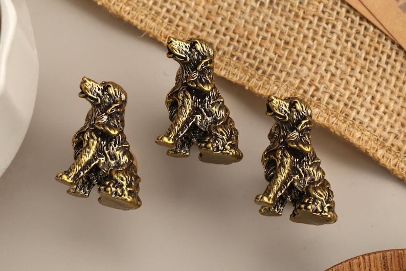 Peut inclure: Trois petites figurines en m&eacute;tal de couleur laiton antique repr&eacute;sentant des chiens assis. Chaque figurine pr&eacute;sente une texture de fourrure d&eacute;taill&eacute;e et une posture avec la t&ecirc;te lev&eacute;e, finie avec une patine sombre et vieillie qui souligne les traits sculpt&eacute;s.