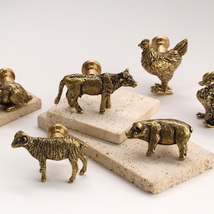 May include: A collection of brass animal-shaped cabinet knobs, including a rabbit, cow, chicken, duck, sheep, and pig. Each knob is mounted on a small, textured, rectangular stone. The knobs are detailed and have a vintage aesthetic.