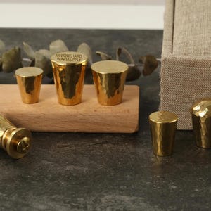 Puede incluir: Una colección de tiradores y pomos de latón martillado de varias formas y tamaños. Los pomos se muestran sobre una tabla de madera y una superficie oscura. El texto "UNIQUEHARD WARESUPPLY" es visible en uno de los pomos.