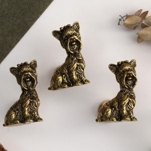 Peut inclure: Un ensemble de quatre boutons de placard en m&eacute;tal couleur laiton antique, chacun en forme de Yorkshire Terrier assis avec une texture de poils d&eacute;taill&eacute;e, des oreilles pointues et un petit n&oelig;ud sur la t&ecirc;te, dot&eacute; d'une base circulaire robuste.