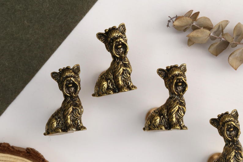 Peut inclure: Un ensemble de quatre boutons de placard en m&eacute;tal couleur laiton antique, chacun en forme de Yorkshire Terrier assis avec une texture de poils d&eacute;taill&eacute;e, des oreilles pointues et un petit n&oelig;ud sur la t&ecirc;te, dot&eacute; d'une base circulaire robuste.