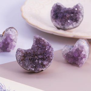 以下が含まれることがあります： 紫色の色合いのアメジストクリスタルクラスター。白いお皿とピンクの表面に配置されています。1つは三日月型で、「UNIQUEHARD WARESUPPLY」の文字が見えます。他のクラスターはハート型です。