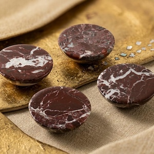 May include: Four circular cabinet knobs made of polished dark maroon stone with prominent white veining. Each knob features a smooth, rounded top surface and a deep red color palette with natural mineral patterns throughout the stone material.