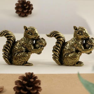 Puede incluir: Dos figuras de ardillas de color bronce, cada una sosteniendo una nuez. Las ardillas tienen pelaje detallado y colas rizadas. Piñas y un trozo de madera están dispersos, sugiriendo un tema de bosque.