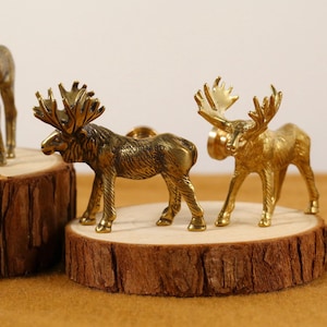 May include: Bronze-colored moose figurines with detailed antlers stand on wooden slices. The figurines have a vintage aesthetic and are likely decorative items. The text "UNIQUEHARD WARESUPPLY" is visible on one of the moose.