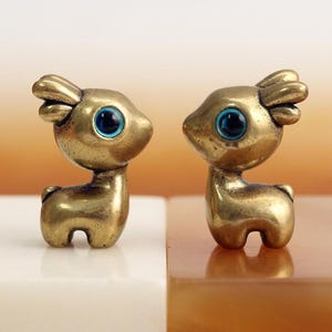 Puede incluir: Dos pequeñas figuras de ciervo de latón estilizadas frente a frente. Cada ciervo tiene un cuerpo liso de color dorado metálico con una cabeza redondeada, orejas cortas y un ojo circular prominente con un iris de color azul brillante.