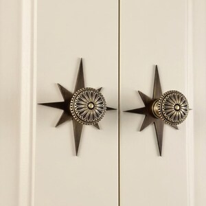 May include: Two antique brass cabinet knobs mounted on a cream-coloured surface. Each knob features a circular, floral-patterned face centred on a multi-pointed star-shaped backplate with a dark, weathered metallic finish and sharp, elongated rays.